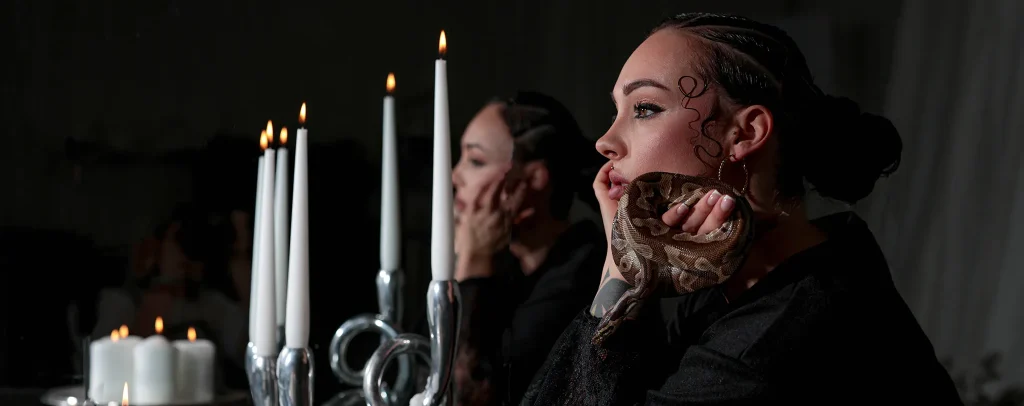 Gothic scene: a woman in black with dramatic makeup gazes at lit candles in a silver candelabrum.