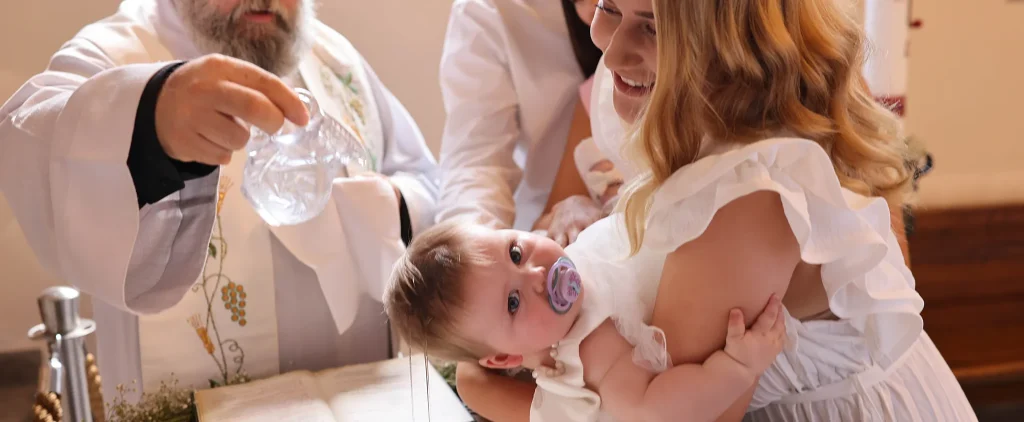 Priest pours water over a baby during a baptism as a smiling woman holds the infant and others look on in a church setting.