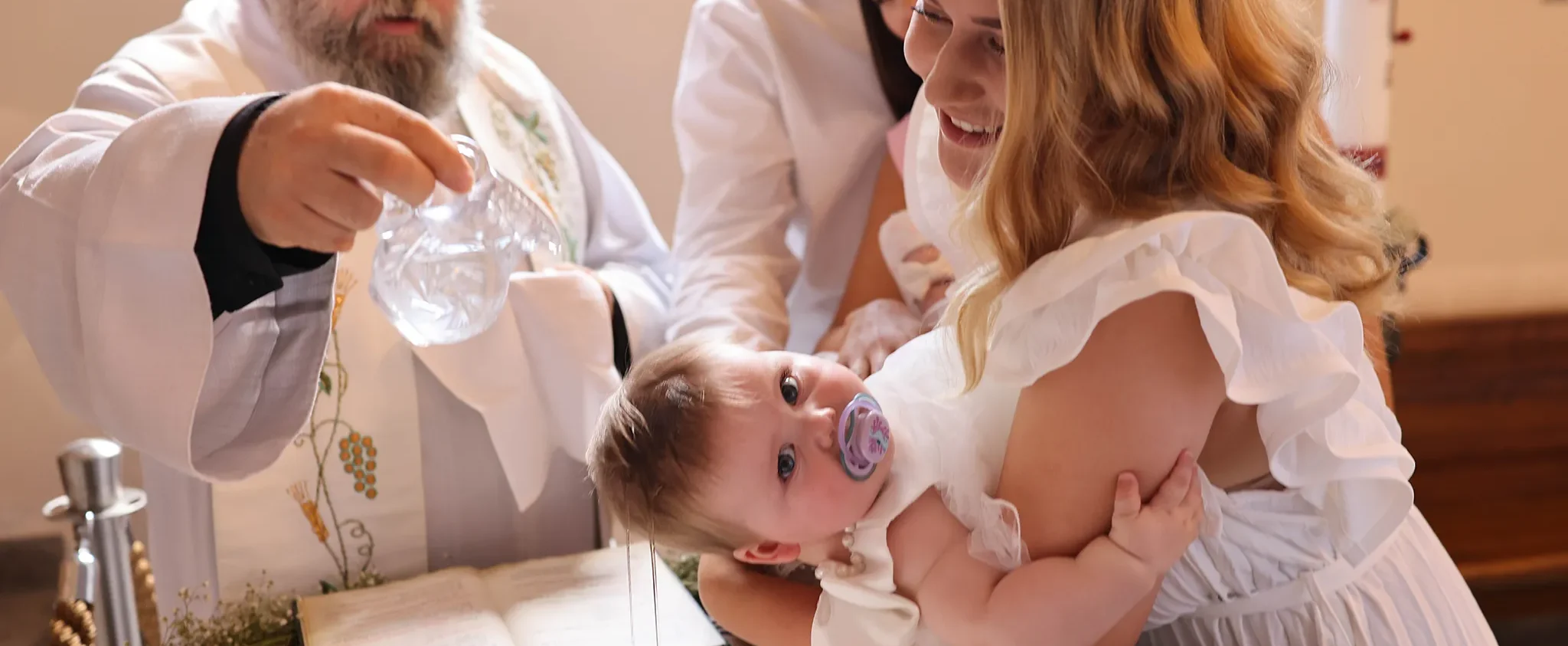 Priest pours water over a baby during a baptism as a smiling woman holds the infant and others look on in a church setting.