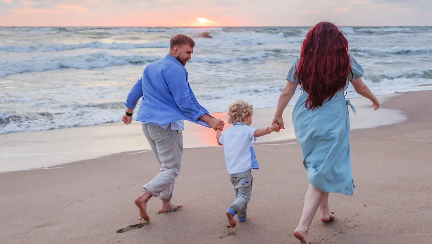 A family of three holding hands and running along the wet shoreline at sunset with waves behind them.