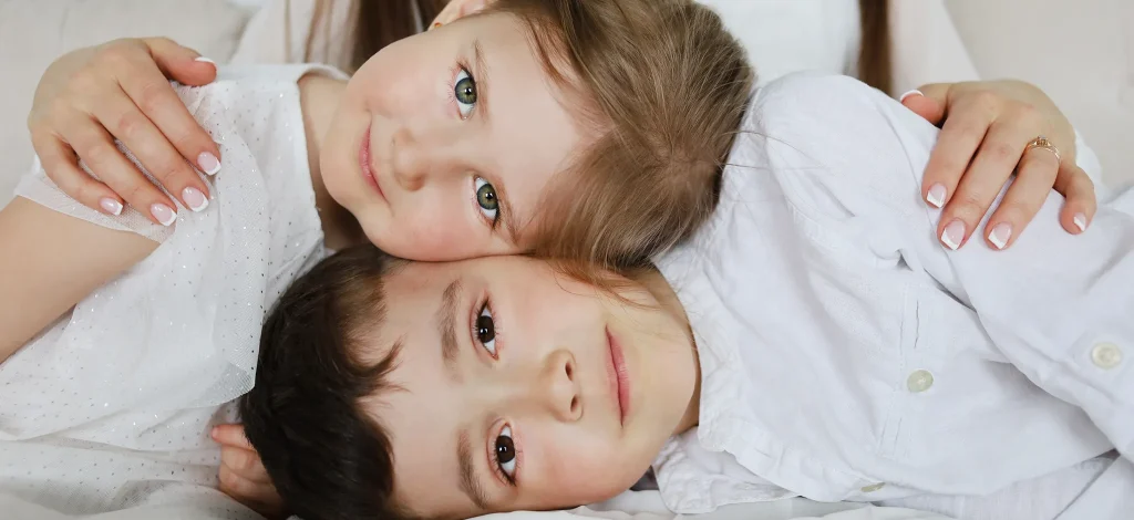 Three children cuddled together on a bed, heads close and smiling at the camera (sisters/brothers).