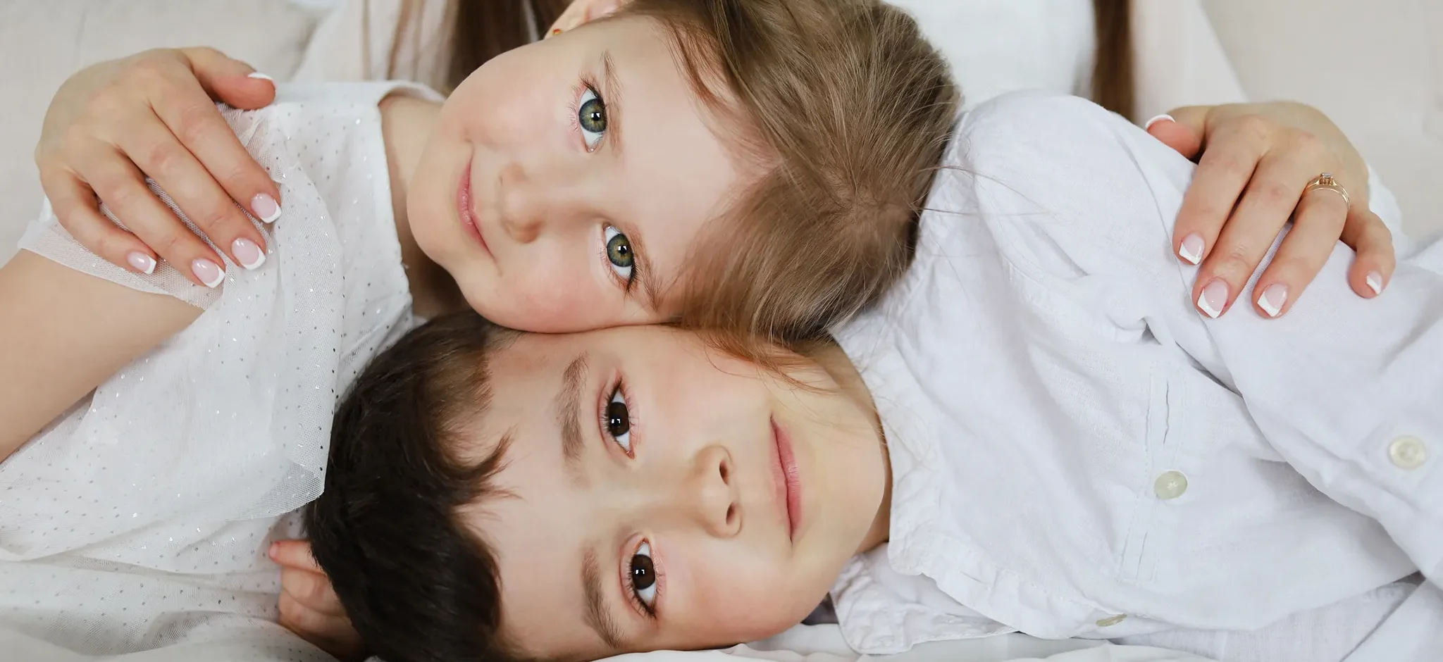 Three children cuddled together on a bed, heads close and smiling at the camera (sisters/brothers).