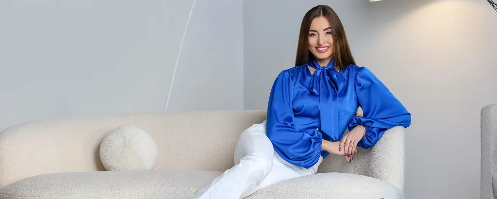 Smiling woman in a royal blue blouse and white pants sits on a beige sofa in a modern living room, facing the camera.
