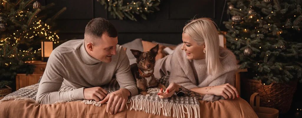 Couple and their dog relaxing on a bed in a festively lit living room with Christmas trees and wreaths nearby (cozy holiday scene).